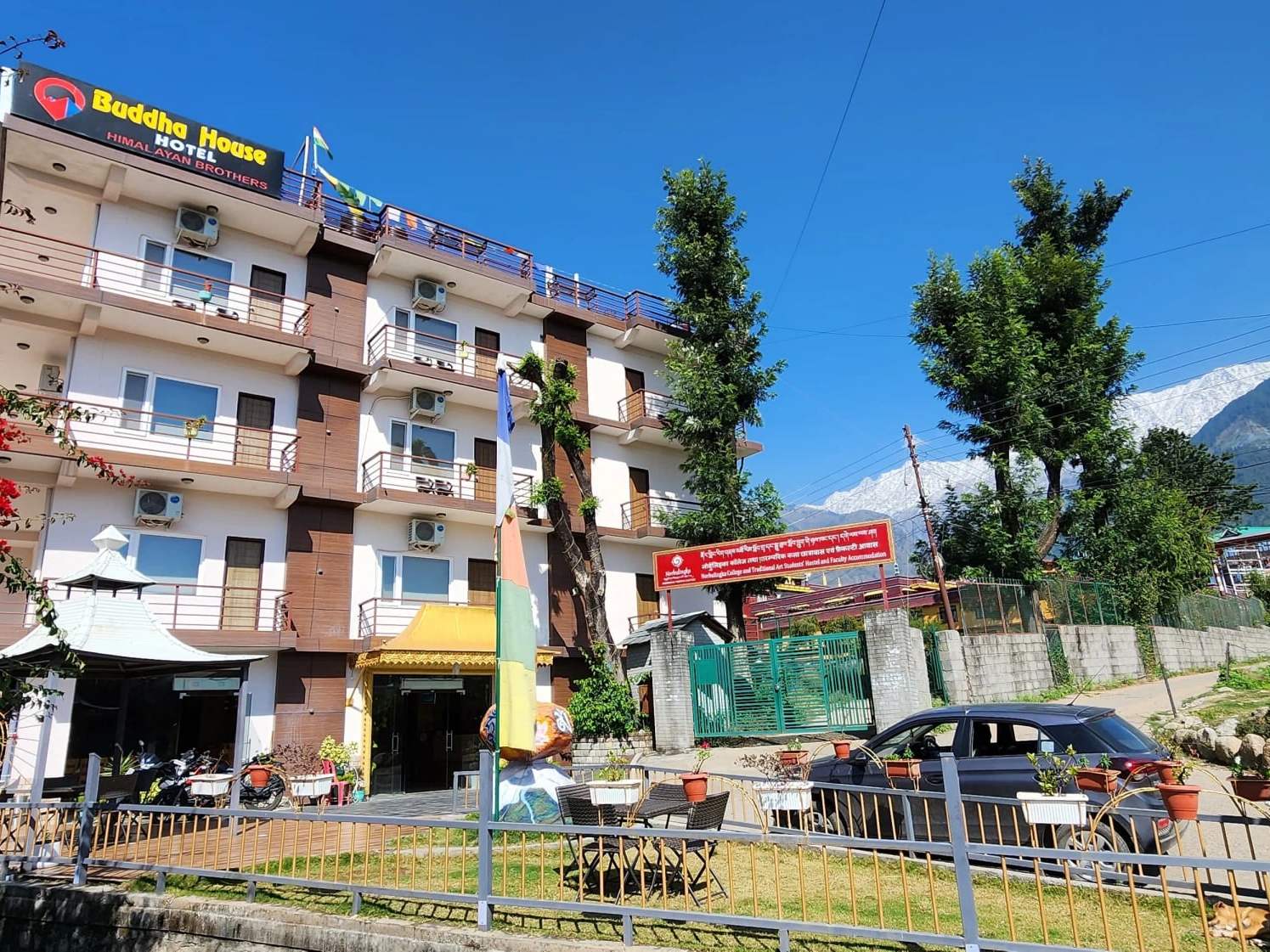 Hotel Buddha House: a five-storey building with balconies and its rooftop sign, a garden in front and snow on the Dhauladhar behind